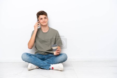 Photo for Teenager Russian man sitting on the floor isolated on white background holding coffee to take away and a mobile - Royalty Free Image