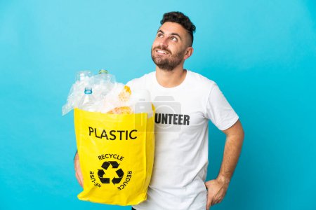 Téléchargez les photos : Homme caucasien tenant un sac rempli de bouteilles en plastique pour recycler isolé sur fond bleu penser une idée tout en levant les yeux - en image libre de droit