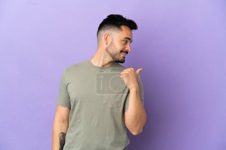 Téléchargez les photos : Jeune homme caucasien isolé sur fond violet pointant vers le côté pour présenter un produit - en image libre de droit