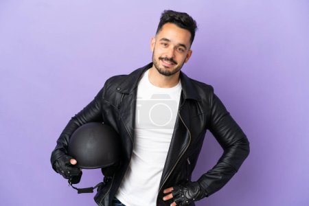 Téléchargez les photos : Jeune homme avec un casque de moto isolé sur fond violet posant avec les bras à la hanche et souriant - en image libre de droit