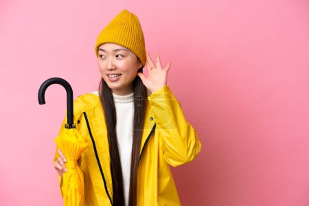 Téléchargez les photos : Jeune femme chinoise avec manteau imperméable et parapluie isolé sur fond rose écoutant quelque chose en mettant la main sur l'oreille - en image libre de droit
