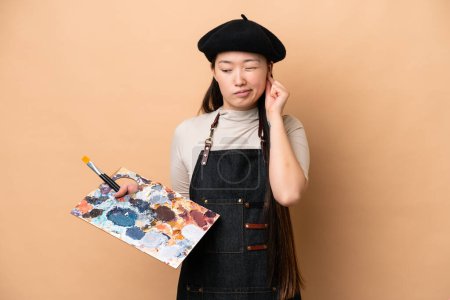 Foto de Joven pintora china aislada sobre fondo beige frustrada y cubriendo oídos - Imagen libre de derechos