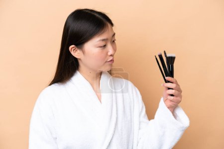 Photo for Young Chinese woman isolated on beige background holding makeup brush and lookin it - Royalty Free Image