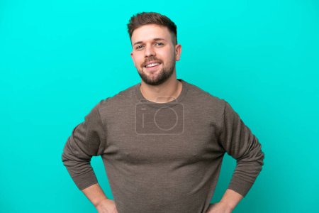 Photo for Young caucasian man isolated on blue background posing with arms at hip and smiling - Royalty Free Image