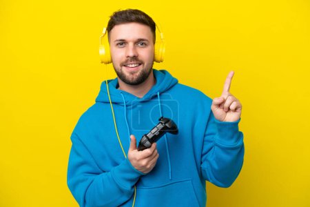 Photo for Young caucasian man playing with a video game controller isolated on yellow background showing and lifting a finger in sign of the best - Royalty Free Image