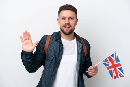 Foto de Joven hombre caucásico sosteniendo una bandera del Reino Unido aislado sobre fondo blanco saludando con la mano con expresión feliz - Imagen libre de derechos