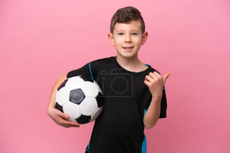 Téléchargez les photos : Petit joueur de football caucasien garçon isolé sur fond rose pointant vers le côté pour présenter un produit - en image libre de droit