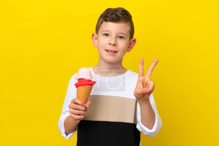 Téléchargez les photos : Petit garçon caucasien avec une crème glacée cornet isolé sur fond jaune souriant et montrant signe de victoire - en image libre de droit