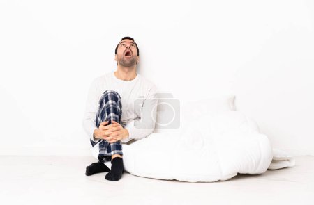 Téléchargez les photos : Homme caucasien en pyjama assis sur le sol à l'intérieur regardant vers le haut et avec expression surprise - en image libre de droit