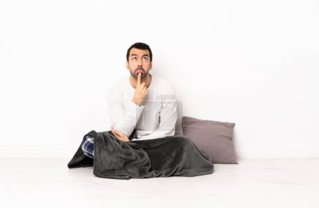 Téléchargez les photos : Homme caucasien en pyjama assis sur le sol à l'intérieur ayant des doutes tout en levant les yeux - en image libre de droit