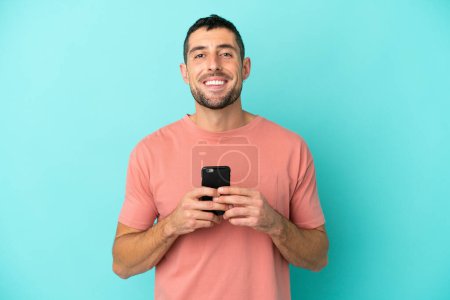 Photo for Young handsome caucasian man isolated on blue background looking at the camera and smiling while using the mobile - Royalty Free Image