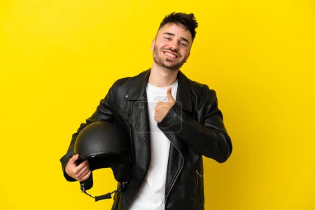 Foto de Joven hombre caucásico con un casco de moto aislado sobre fondo amarillo dando un gesto pulgar hacia arriba - Imagen libre de derechos