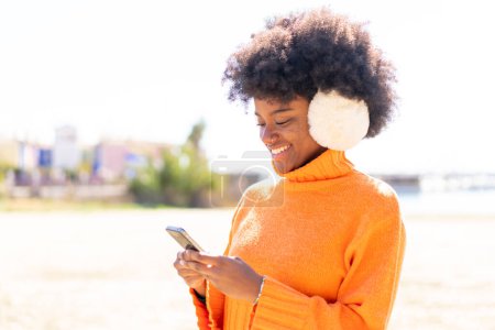 Téléchargez les photos : Afro-américaine fille portant des manchons d'hiver à l'extérieur envoyer un message avec le mobile - en image libre de droit