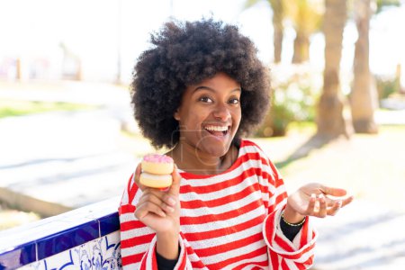 Foto de Chica afroamericana sosteniendo una dona al aire libre con expresión facial impactada - Imagen libre de derechos