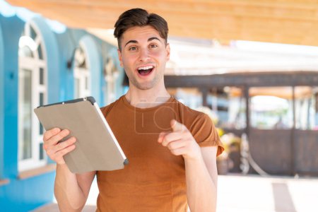 Foto de Joven hombre guapo sosteniendo una tableta al aire libre sorprendido y apuntando hacia delante - Imagen libre de derechos
