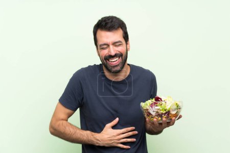 Photo for Young handsome man with salad over isolated green wall smiling a lot - Royalty Free Image