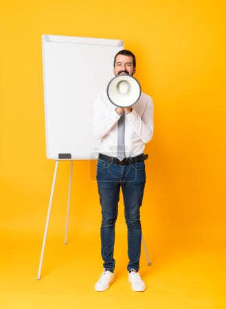 Téléchargez les photos : Plan complet de l'homme d'affaires donnant une présentation sur tableau blanc sur fond jaune isolé criant à travers un mégaphone - en image libre de droit