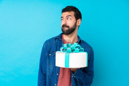 Photo for Young handsome man with a big cake over isolated blue background with confuse face expression - Royalty Free Image