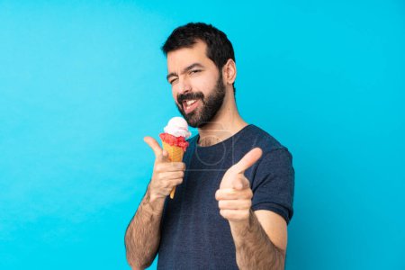 Foto de Joven con un helado de corneta sobre fondo azul aislado apuntando hacia el frente y sonriendo - Imagen libre de derechos