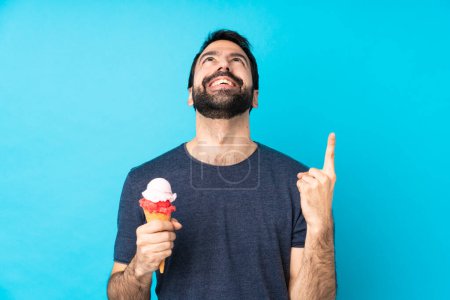 Téléchargez les photos : Jeune homme avec une crème glacée cornet sur fond bleu isolé pointant vers le haut et surpris - en image libre de droit