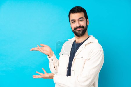 Foto de Joven hombre guapo con chaqueta de pana blanca sobre fondo azul aislado extendiendo las manos a un lado para invitar a venir - Imagen libre de derechos