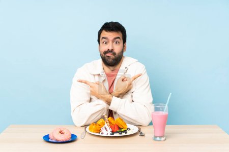Téléchargez les photos : Homme à une table ayant des gaufres pour le petit déjeuner et un milk-shake pointant vers les latéraux ayant des doutes - en image libre de droit