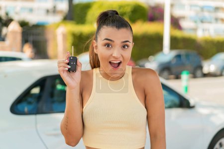 Téléchargez les photos : Jeune jolie femme tenant des clés de voiture à l'extérieur avec surprise et expression faciale choquée - en image libre de droit