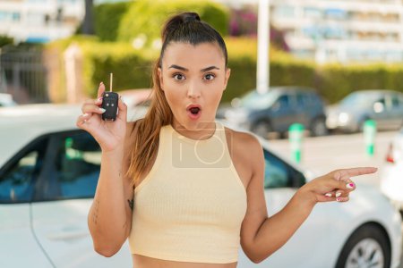 Téléchargez les photos : Jeune jolie femme tenant des clés de voiture à l'extérieur surpris et pointant côté - en image libre de droit