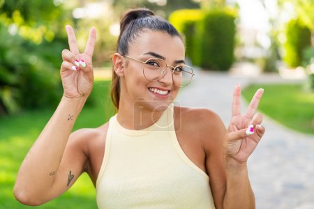 Téléchargez les photos : Jeune jolie femme avec des lunettes à l'extérieur montrant signe de victoire avec les deux mains - en image libre de droit