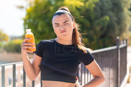 Foto de Joven mujer bastante deportiva sosteniendo un jugo de naranja al aire libre con expresión triste - Imagen libre de derechos