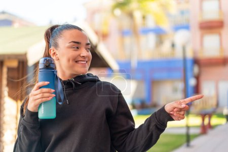 Téléchargez les photos : Jeune jolie femme sportive avec une bouteille d'eau à l'extérieur pointant vers le côté pour présenter un produit - en image libre de droit