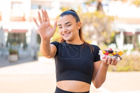 Foto de Joven mujer morena bonita sosteniendo un tazón de fruta al aire libre saludando con la mano con expresión feliz - Imagen libre de derechos