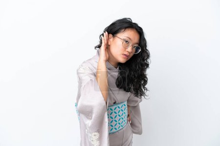 Foto de Joven mujer asiática usando kimono aislado sobre fondo blanco escuchando algo poniendo la mano en la oreja - Imagen libre de derechos