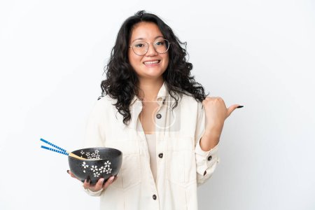Téléchargez les photos : Jeune femme asiatique isolée sur fond blanc pointant vers le côté pour présenter un produit tout en tenant un bol de nouilles avec des baguettes - en image libre de droit