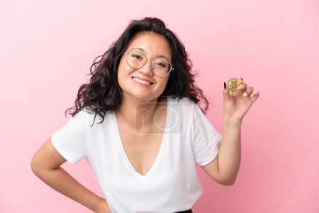 Photo for Young asian woman holding a Bitcoin isolated on pink background posing with arms at hip and smiling - Royalty Free Image