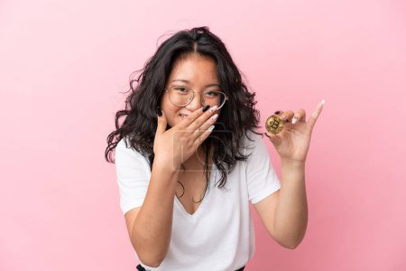 Téléchargez les photos : Jeune femme asiatique tenant un Bitcoin isolé sur fond rose heureux et souriant couvrant la bouche avec la main - en image libre de droit