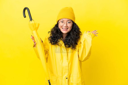Femme asiatique avec manteau imperméable et parapluie isolé sur fond jaune avec expression faciale choquée