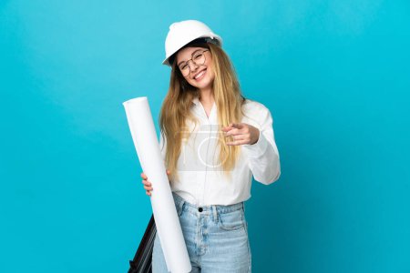 Téléchargez les photos : Jeune architecte femme avec casque et tenant des plans isolés sur fond bleu pointant devant avec expression heureuse - en image libre de droit