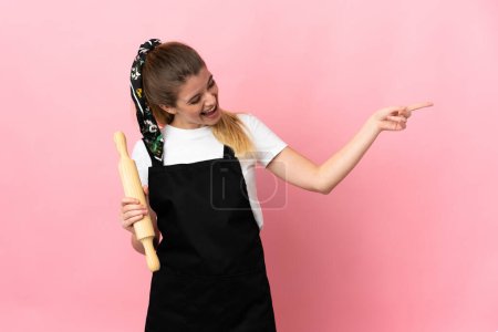 Téléchargez les photos : Jeune femme blonde tenant un rouleau à pâtisserie isolé sur fond rose pointant du doigt vers le côté et présentant un produit - en image libre de droit