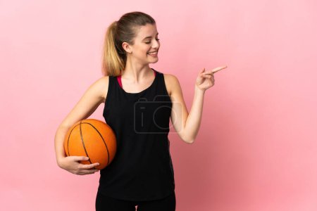 Foto de Joven rubia jugando baloncesto aislada sobre fondo rosa apuntando con el dedo hacia un lado y presentando un producto - Imagen libre de derechos