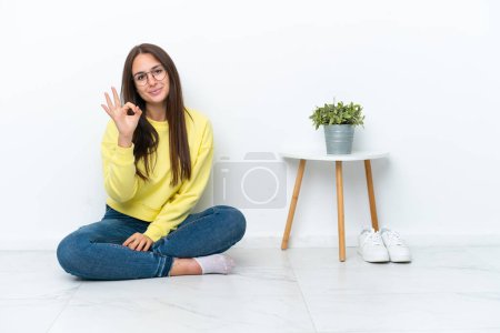 Foto de Mujer ucraniana joven sentada en el suelo de su casa aislada en la pared blanca que muestra signo de ok con los dedos - Imagen libre de derechos