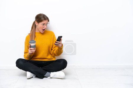 Téléchargez les photos : Jeune femme caucasienne assise sur le sol isolé sur fond blanc tenant du café à emporter et un mobile - en image libre de droit