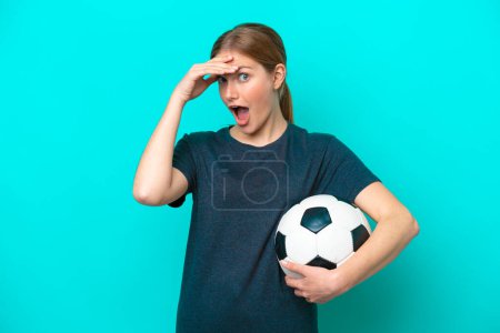 Téléchargez les photos : Jeune joueur de football femme isolée sur fond bleu faisant geste de surprise tout en regardant vers le côté - en image libre de droit