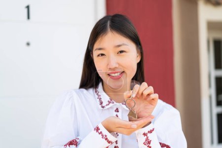 Téléchargez les photos : Jeune femme chinoise à l'extérieur tenant les clés de la maison avec expression heureuse - en image libre de droit