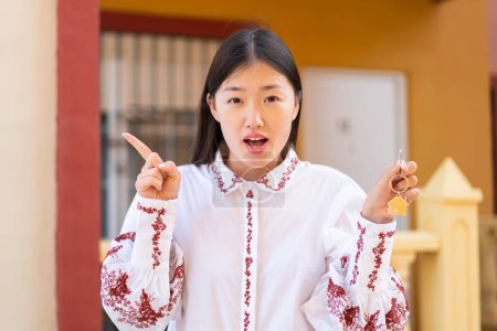 Téléchargez les photos : Jeune femme chinoise tenant les clés de la maison à l'extérieur surpris et pointant côté - en image libre de droit
