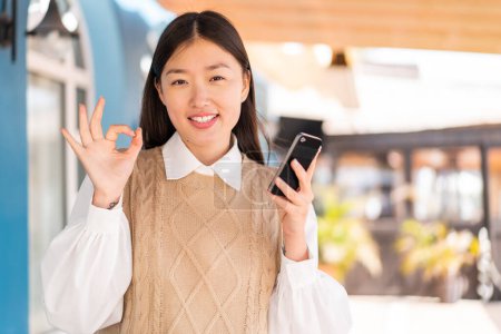 Téléchargez les photos : Jeune femme chinoise à l'extérieur en utilisant un téléphone mobile et faire OK signe - en image libre de droit