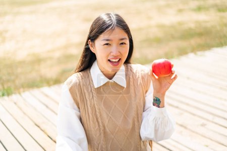 Téléchargez les photos : Jeune femme chinoise avec une pomme à l'extérieur avec une expression faciale surprise et choquée - en image libre de droit