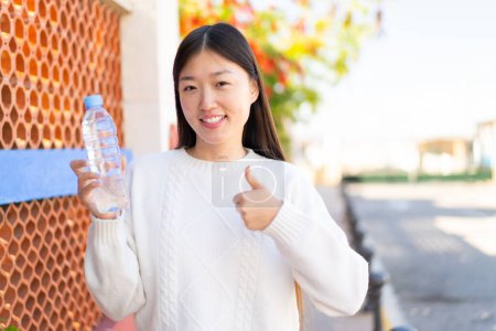 Foto de Mujer china bonita con una botella de agua al aire libre con los pulgares hacia arriba porque algo bueno ha sucedido - Imagen libre de derechos