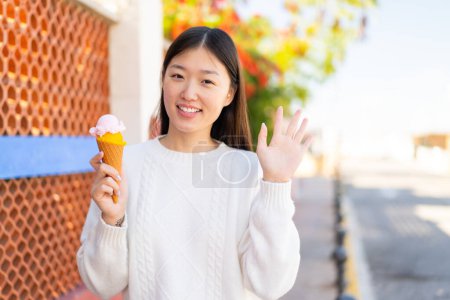 Foto de Mujer china bonita con un helado de corneta al aire libre saludando con la mano con expresión feliz - Imagen libre de derechos