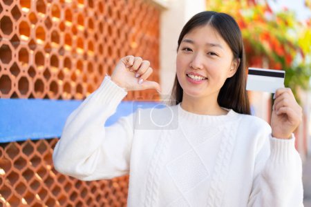 Foto de Bonita mujer china sosteniendo una tarjeta de crédito al aire libre orgullosa y satisfecha - Imagen libre de derechos